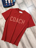 Red t-shirt with 'COACH' printed on it, lying on a wooden floor.