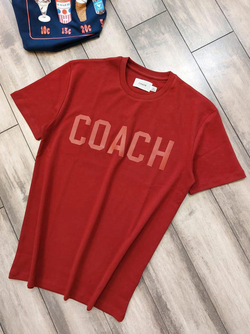 Red t-shirt with 'COACH' printed on it, lying on a wooden floor.